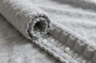 A high-quality close-up shot of a cozy, textured grey weighted blanket, partially unrolled to reveal the inner grid structure. Inside the small square pockets, smooth white glass beads are visible, showing the internal distribution of weight. The lighting is soft and natural, highlighting the fabric texture and the glass beads.