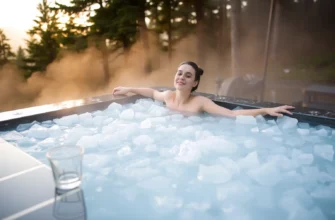 A serene scene of a person standing in a clear cold plunge tub filled with icy water, surrounded by a natural forest setting at sunrise. The person is relaxed, wearing minimal clothing, with a small glass of water placed beside the tub. The background includes misty trees and soft light, conveying calm and rejuvenation.