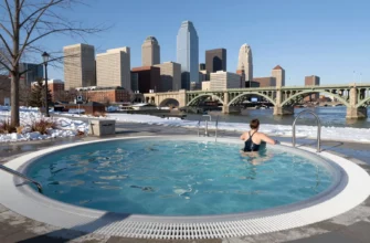 A realistic outdoor cold plunge pool in Minneapolis, featuring a shallow pool of icy water surrounded by winter scenery, a person in a swimsuit standing at the edge, and the Minneapolis skyline or a recognizable landmark such as the Stone Arch Bridge in the background, all under a clear blue sky.