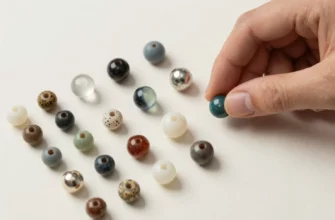 A close-up, high-resolution image of a variety of weighted blanket beads arranged neatly on a clean, neutral-colored surface. The beads should include different sizes, colors, and textures (e.g., glass, plastic, metal, and silicone). A hand (no face or body visible) is gently picking up one bead, with a small, open guidebook or reference sheet beside the beads, but without any visible text or lettering. The lighting should highlight the beads’ weight and texture, creating subtle shadows for depth. The background should be simple and uncluttered, focusing attention on the beads and the hand.