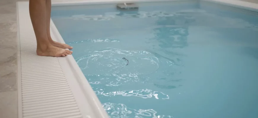 A person standing at the edge of a hot tub with steam rising, transitioning to a cold plunge pool with clear blue water, both pools side by side in a serene spa setting, with natural lighting and subtle reflections