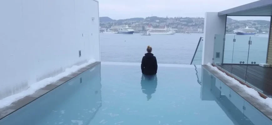 A person standing in a clear, icy plunge pool surrounded by minimalist Scandinavian architecture, with a view of the Copenhagen harbor in the background, reflecting the cold water and crisp atmosphere.