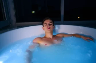 A serene nighttime scene of a person standing in a clear, cold plunge tub surrounded by soft, cool lighting. The tub is filled with sparkling, icy water, and the person is relaxed, with a calm expression, suggesting preparation for sleep. The background is minimal, featuring subtle hints of a dark, starry sky and a faint glow from a nearby window, creating a tranquil, restful atmosphere.