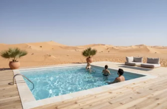A serene desert wellness studio set against a backdrop of golden sand dunes under a clear blue sky, featuring a sleek modern cold plunge pool with crystal‑clear water, surrounded by minimalist wooden decking and lush green potted plants. A few relaxed individuals in swimwear are gently stepping into the pool, while a yoga mat and a small meditation corner with cushions are visible nearby. The scene captures the contrast between the hot desert environment and the refreshing coolness of the plunge