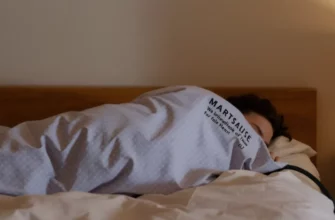 A calm bedroom scene featuring a person peacefully sleeping on a bed, covered by a soft weighted blanket. The setting includes subtle medical elements such as a faint heart rate monitor display or a gentle glow around the blanket to suggest safety and comfort. The lighting is warm and soothing, with no visible text or symbols that could be interpreted as instructions or branding.
