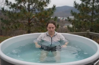 A person standing in a clear cold plunge tub filled with icy water, surrounded by a scenic Knoxville outdoor setting with pine trees and a hint of the Great Smoky Mountains in the background, wearing a light jacket, looking relaxed