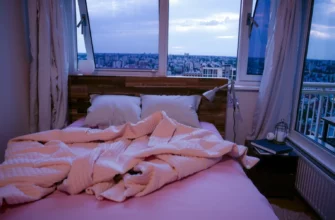 A cozy bedroom scene featuring a neatly made bed with a soft, textured weighted blanket draped over it, a wooden nightstand with a small lamp and a book, and a window showing a calm evening sky outside