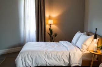 A serene, softly lit bedroom interior featuring a plush, low-profile bed with crisp white linens, a small wooden nightstand holding a single candle, a gently draped sheer curtain, and a subtle, warm glow from a bedside lamp. The room should convey calmness and tranquility, with muted earth tones and natural textures such as a woven rug and a potted plant. No visible furniture or décor beyond the described elements, and no people or text in the scene.