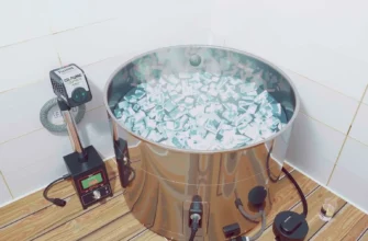 A modern indoor cold plunge setup featuring a sleek stainless steel tub filled with crystal-clear icy water, a compact electric heater mounted beside the tub, a visible water circulation pump with transparent tubing, and a control panel with digital gauges (no text visible). The scene includes subtle steam rising from the water, soft ambient lighting highlighting the equipment, and a clean minimalist bathroom environment with tiled walls and a wooden deck floor.