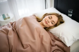 A person comfortably lying on a bed covered with a weighted blanket, looking relaxed and peaceful. The room is softly lit, with a focus on the blanket's texture and the person's serene expression. The background includes subtle elements like a bedside table with a book and a glass of water, suggesting a calming environment.