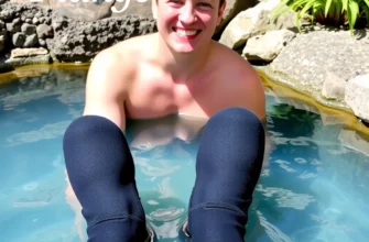 A person wearing neoprene booties is standing in a cold plunge pool. The person is smiling and appears comfortable. The pool is surrounded by rocks and plants. The lighting is bright and natural.