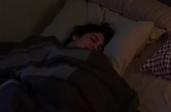 A person lying comfortably on a bed, wrapped in a weighted blanket. The room is dimly lit and cozy, with soft textures visible. Focus on the feeling of relaxation and security.