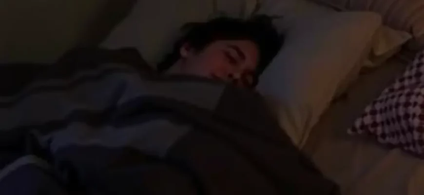 A person lying comfortably on a bed, wrapped in a weighted blanket. The room is dimly lit and cozy, with soft textures visible. Focus on the feeling of relaxation and security.