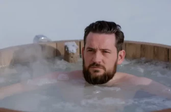 A man with a beard is immersed in a cold plunge tub outdoors. The tub is made of wood and is surrounded by a snowy landscape. Steam is rising from the water. He has a look of determination on his face. Focus on the man and the tub.