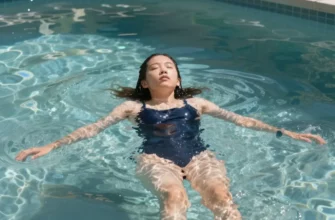 A person taking a cold plunge in a clear, outdoor pool on a sunny day. The person is wearing a swimsuit and appears refreshed and invigorated. The water is crystal clear, and sunlight reflects off the surface. Lush green plants surround the pool.
