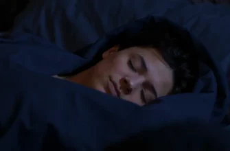 A person sleeping peacefully under a chain weighted blanket in a dimly lit bedroom. The focus is on the blanket's texture and the person's relaxed posture. Soft, diffused lighting creates a calming atmosphere.