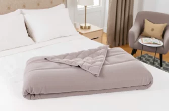 A cozy bedroom scene with a neatly made bed featuring a high-quality weighted blanket in a neutral color, such as gray or beige. The blanket should be folded at the foot of the bed, showcasing its texture and weight. The room should have soft lighting, a comfortable chair, and a small side table with a book and a cup of tea, creating a relaxing atmosphere.