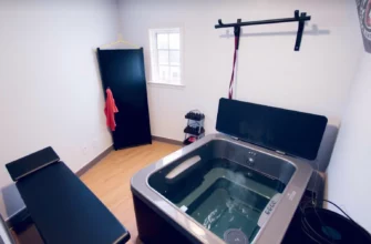A compact home gym setup featuring a cold plunge tub placed neatly in a corner, surrounded by minimal fitness equipment like a foldable bench, resistance bands, and a wall-mounted pull-up bar. The space is well-lit with natural light from a small window, showing efficient use of limited square footage. The tub is modern, made of sleek black or dark gray composite material with a simple lid, filled with clear water and subtle steam rising slightly to indicate cold temperature. No text, signs, or