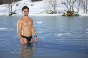 A person standing in cold water up to their waist, outdoors in a natural setting like a lake or icy pool, with a calm and determined expression, wearing minimal clothing like swim trunks or shorts, surrounded by a cold, wintry environment with bare trees or snow in the background, sunlight reflecting off the water surface