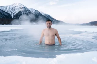 A person standing in icy water with steam rising from their body, surrounded by snow-covered mountains under a pale winter sky, showing a moment of cold exposure therapy in a natural setting